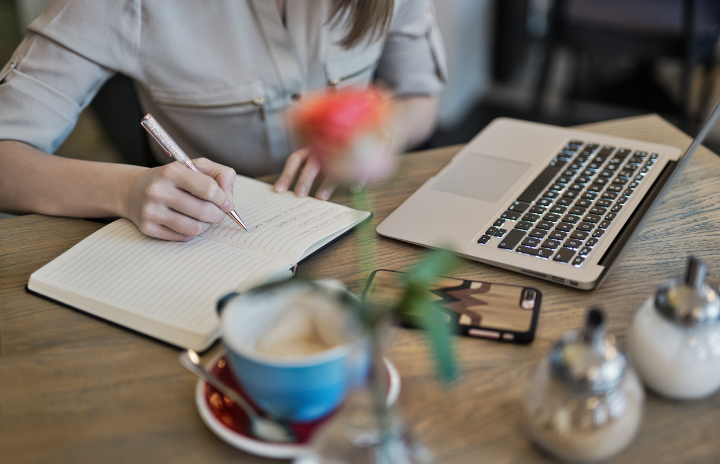 A girl writing on a notebook with a laptop and a cup of coffee